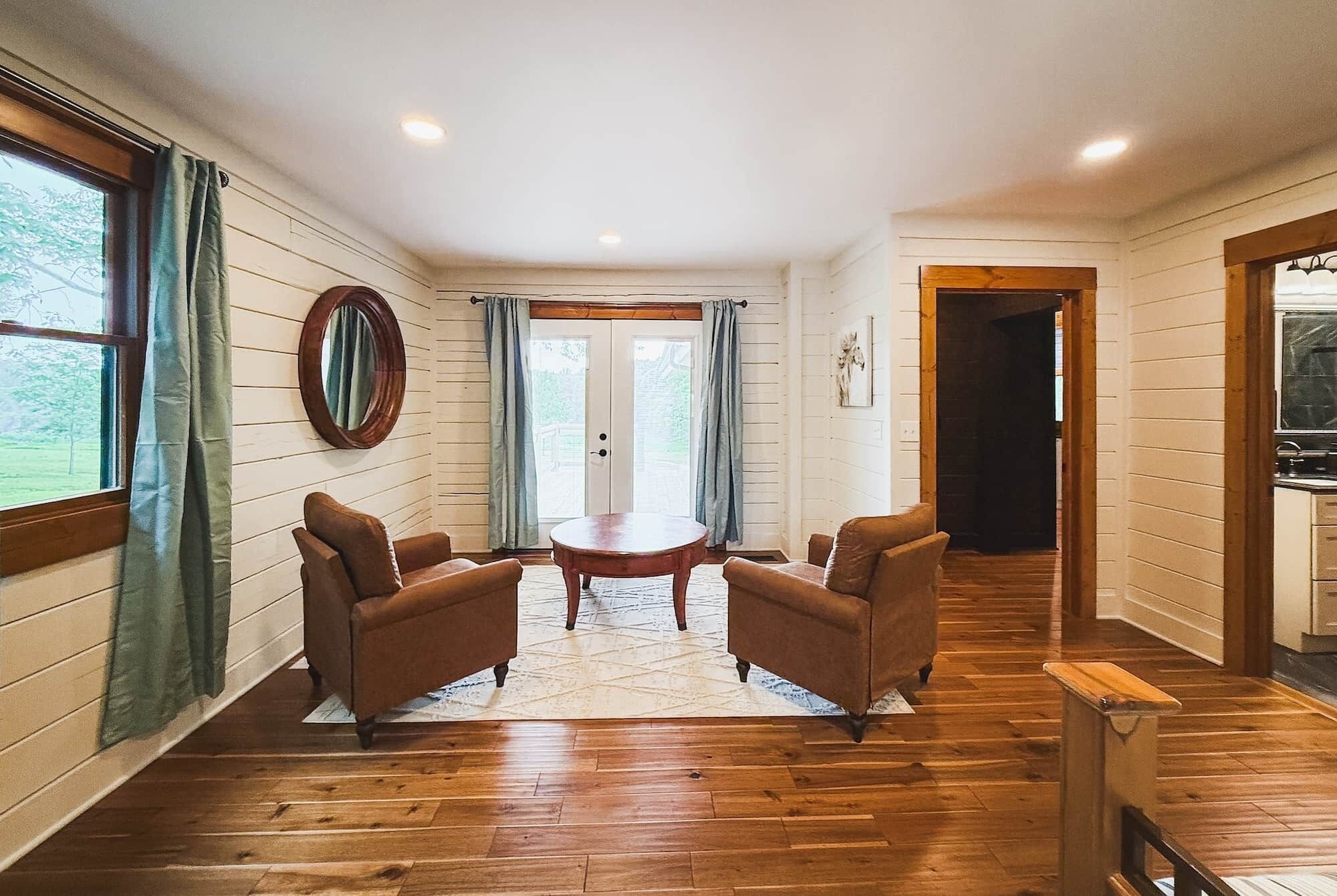 A room with a wooden floor, a round table, two armchairs, a mirror, and a door leading to another room.