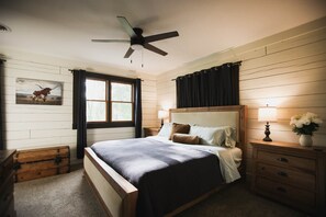 A bedroom with a bed, a nightstand, a lamp, a ceiling fan, a window with curtains, and a painting on the wall. - Dutton Chapel Lodge (Golconda No. 2 Precinct)