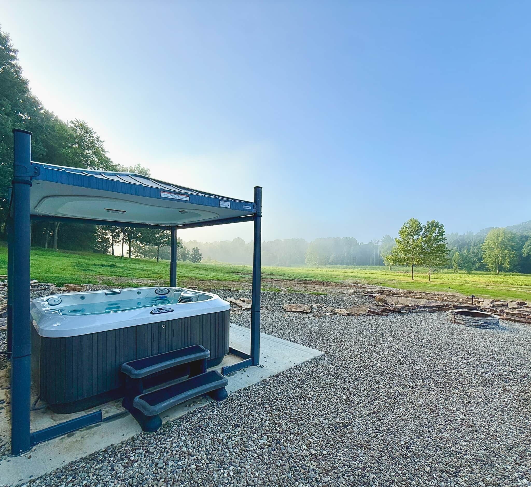 A hot tub under a covered structure with a gravel area and a view of a green landscape.