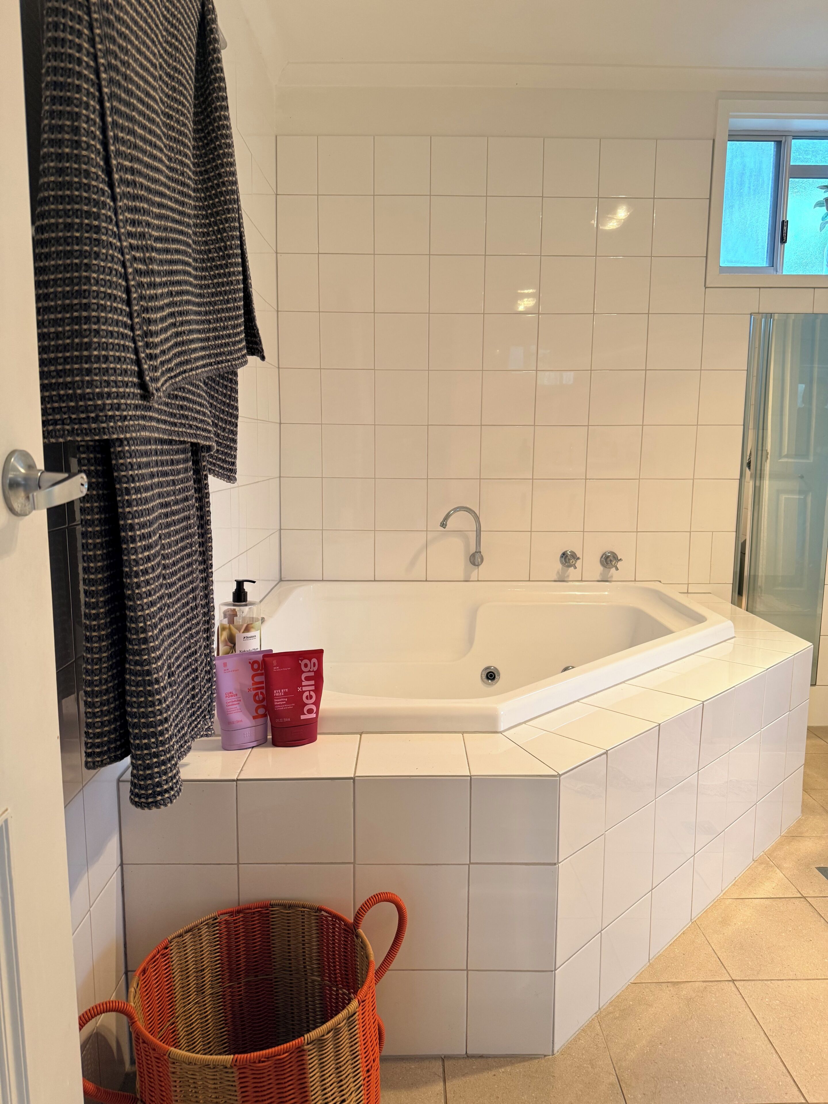 A bathroom with a white bathtub, a towel rack with a towel, a bottle of shampoo, a red cup, and a wicker basket.