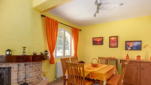 Ein Essbereich mit Kamin aus Ziegelsteinen, Holzsesseln, einem Tisch mit gelber Tischdecke und einem Fenster mit orangenen Vorhängen.