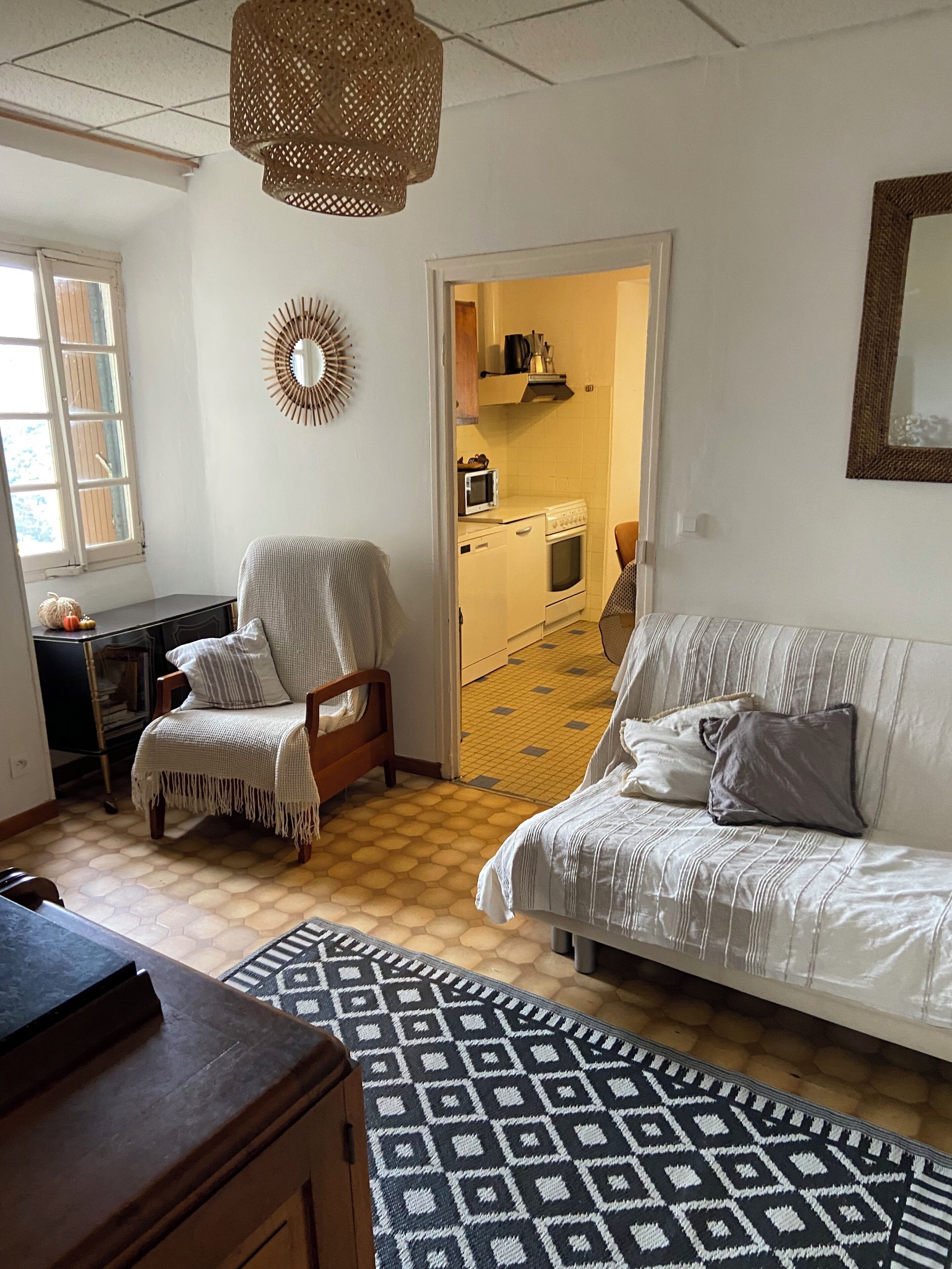 Un salon chaleureux avec un canapé, un fauteuil et un tapis à motifs. On aperçoit un coin cuisine à travers une porte ouverte.