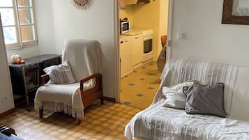 A cozy living room with a sofa, armchair, and a patterned rug. A kitchen area is visible through an open doorway.