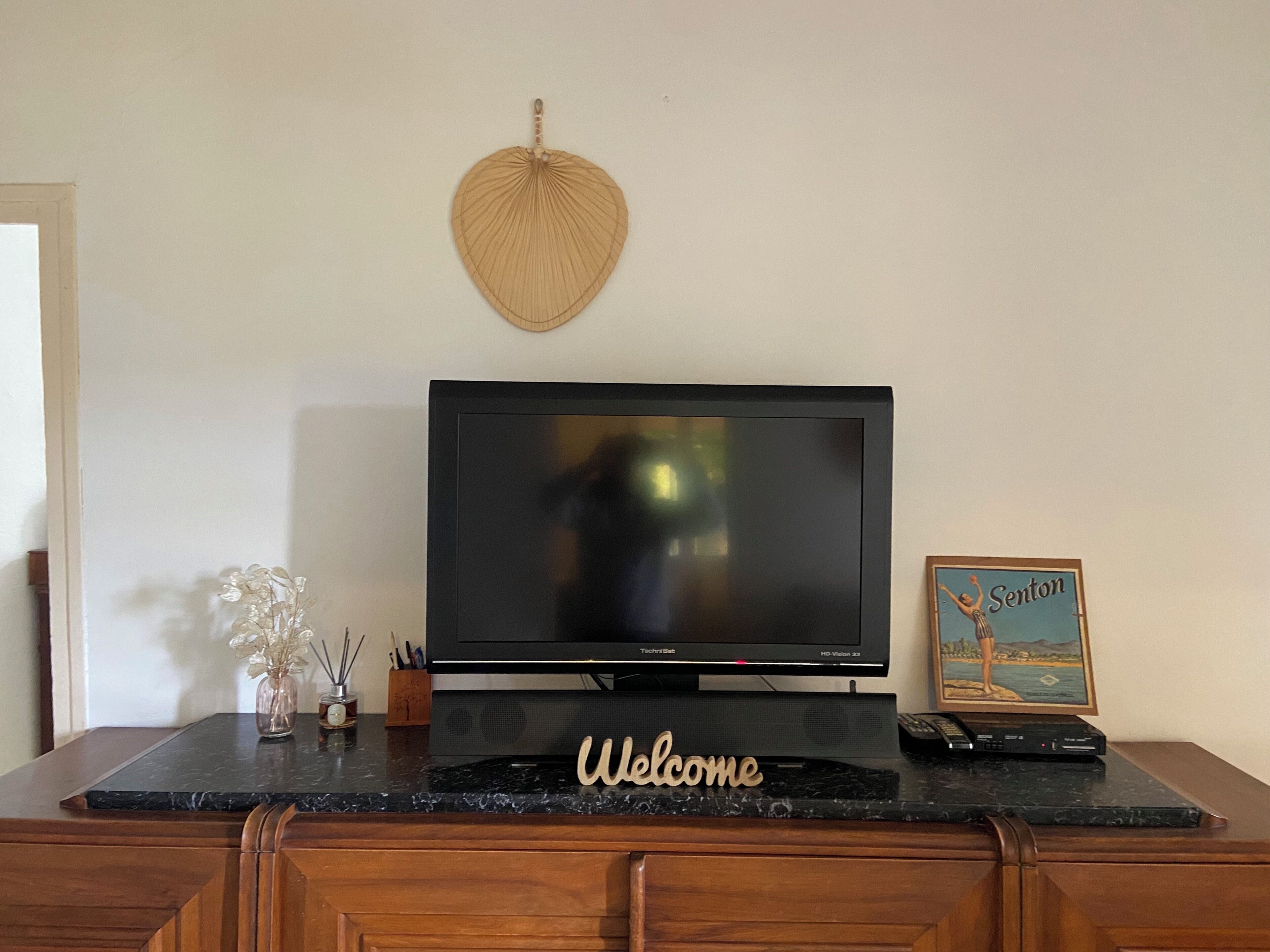 A flat-screen TV on a black stand with a 'Welcome' sign, a framed picture, and a vase with decorative items on a marble countertop.