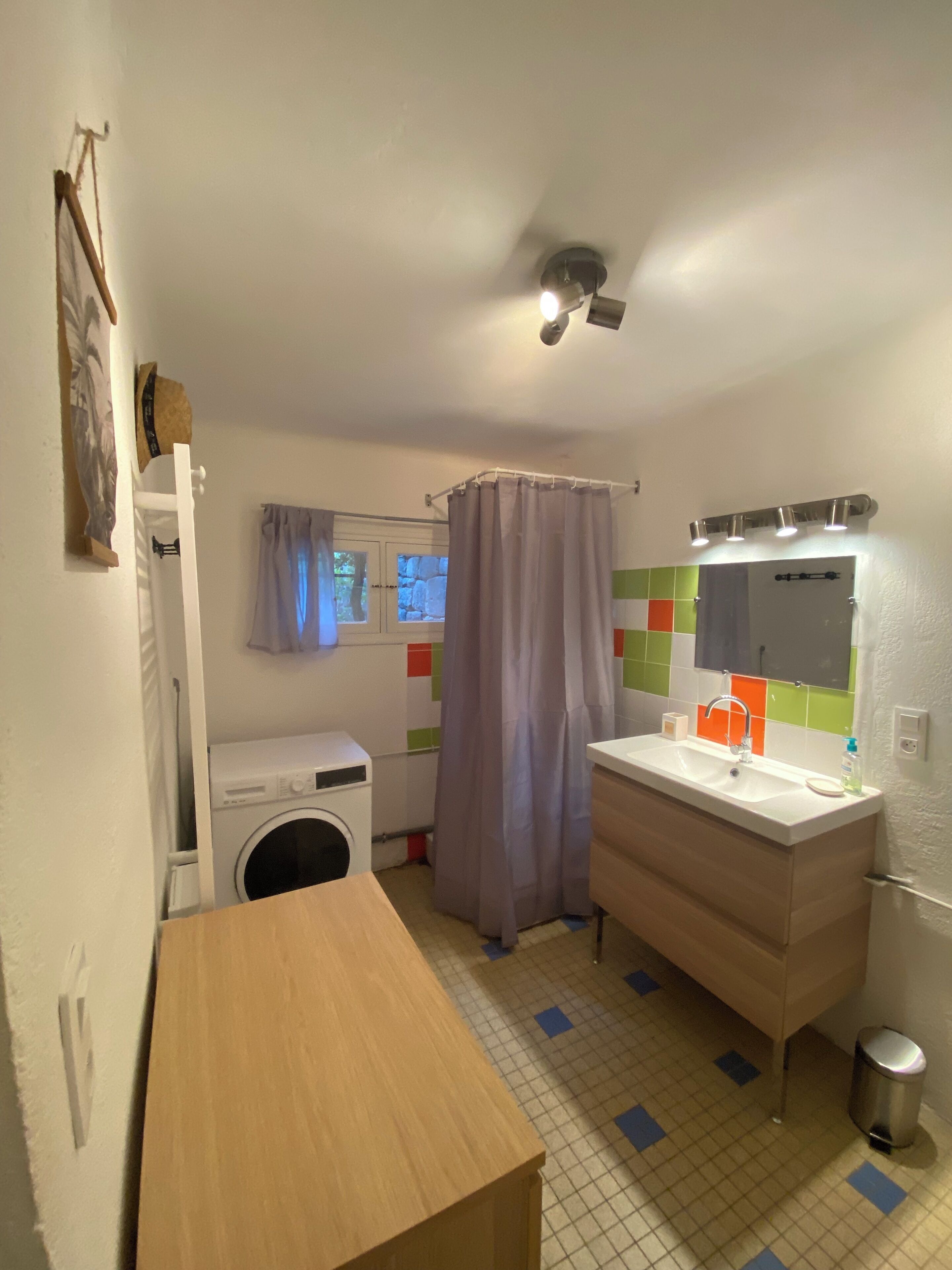 Een kleine badkamer met een wasmachine, een wastafel met spiegel, een douchegordijn en een kleine prullenbak.