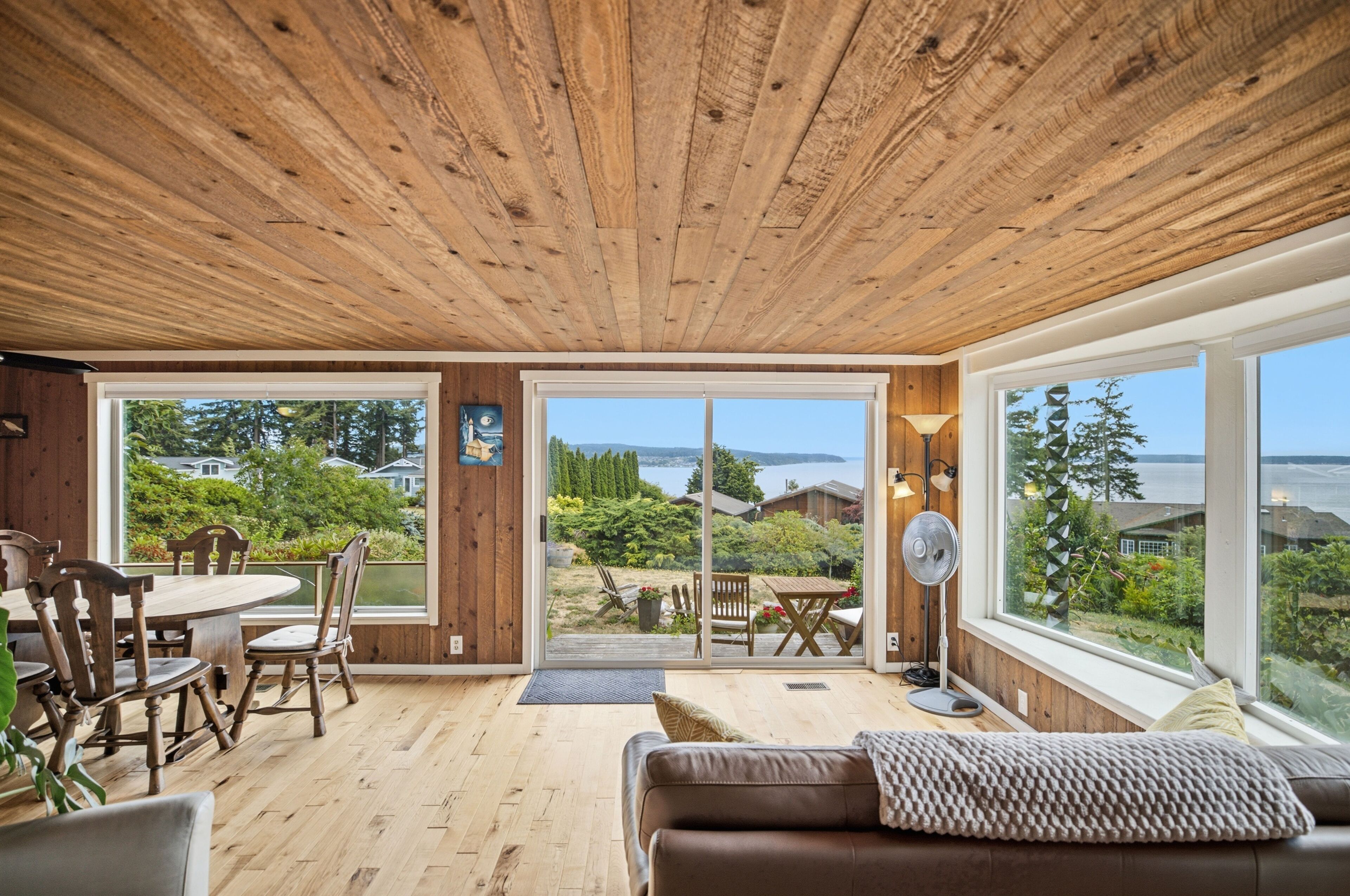 Un plafond en bois, un coin repas avec des chaises et une table, un canapé et de grandes fenêtres donnant sur un paysage verdoyant et un plan d’eau.