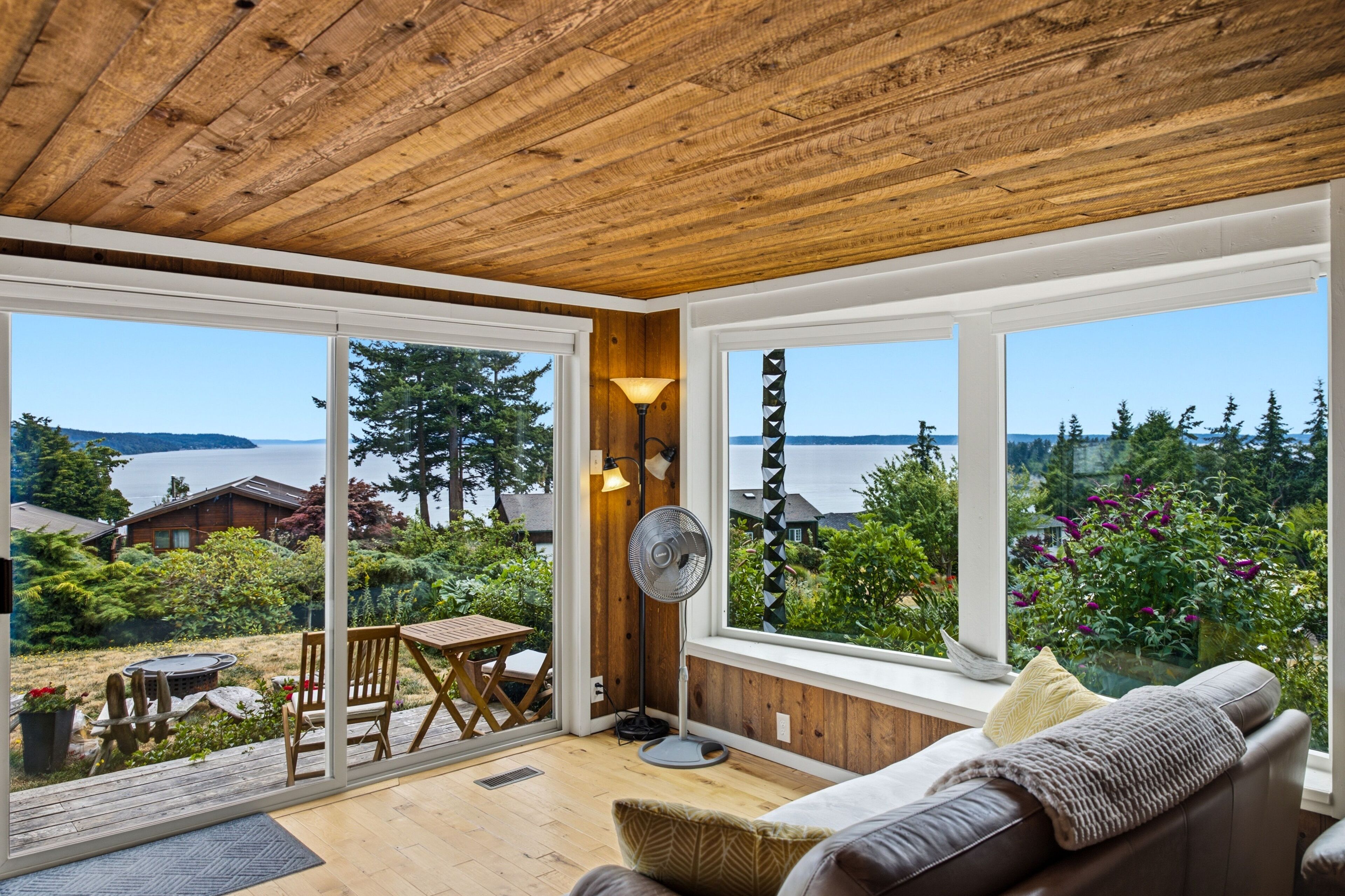 Un interior moderno con techo de madera, grandes ventanales que ofrecen vista a un lago y un bosque, una zona de comedor con mesa y sillas, y un rincón de estar acogedor con sofá.