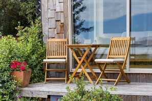 Eine Holzterrasse mit zwei Holzstühlen und einem Tisch, umgeben von Grünpflanzen und einem großen Fenster.