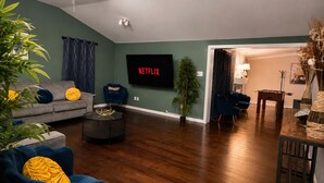 Uma sala de estar com sofá, poltronas, mesa de centro, televisão e uma área de jantar com mesa e cadeiras.