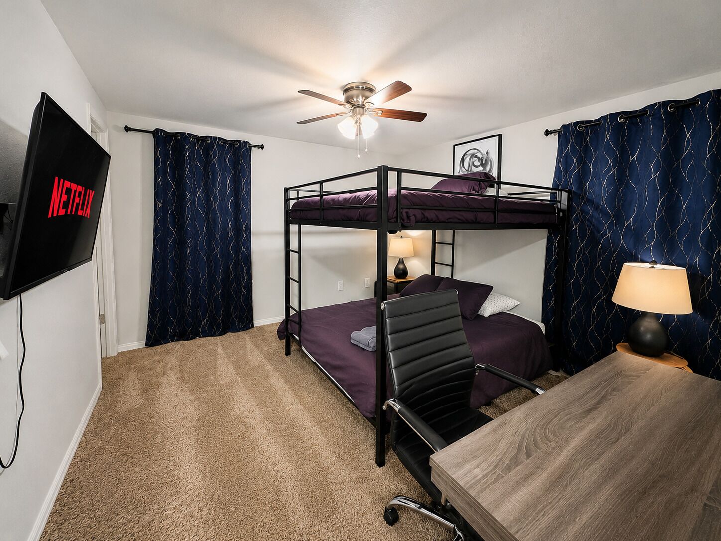 A bunk bed room with a ceiling fan, a TV mounted on the wall, and a desk with a lamp.