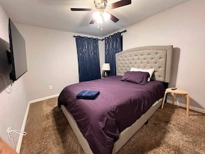A bedroom with a bed, a ceiling fan, a nightstand, and a wall-mounted TV.