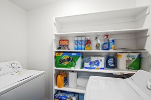 A laundry room with a washing machine, shelves stocked with cleaning supplies, and paper towels.