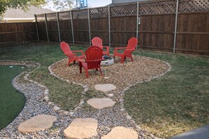 A backyard with a small putting green, red Adirondack chairs, a fire pit, and a wooden fence. - Spacious home with putting green and game room (Carrollton)