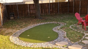 Un jardín trasero con un campo de prácticas de golf, una valla de madera y sillas rojas.