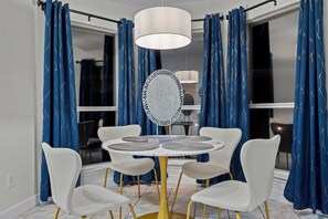 A dining area with a round table, white chairs, and a central mirror. Blue curtains with a pattern are on the windows. - Frisco 4BR | Crew, Team & Relocation Ready (Frisco)