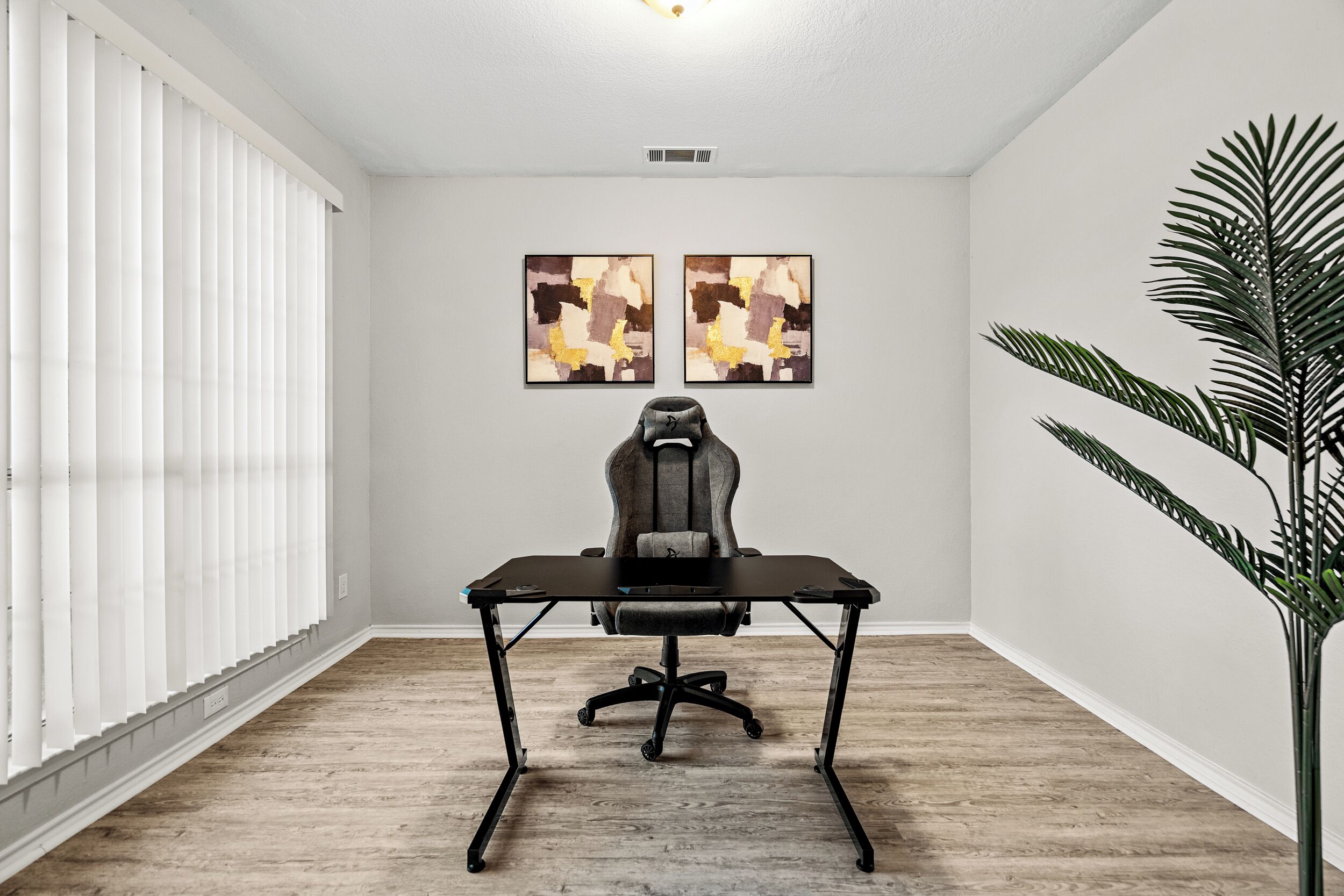 Une pièce comprenant un fauteuil de jeu, un bureau, deux tableaux abstraits, une grande plante et une fenêtre avec des stores.