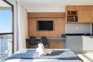 A hotel room with a bed, a desk with two chairs, a flat-screen TV, a kitchenette, and a large window. - Studio in Condo. w/ Sea View & Pool Rgf0508 (Salvador)