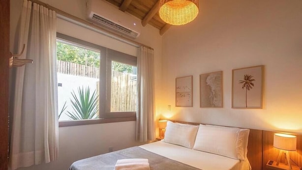 A hotel room with a bed, two bedside lamps, a window with curtains, and wall art. - Studio w/ Balcony in Arraial D'ajuda Ipe0001 (Porto Seguro)