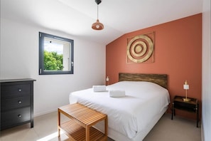 A bedroom with a bed, a wooden nightstand, a dresser, a window with a view, and a pendant light.