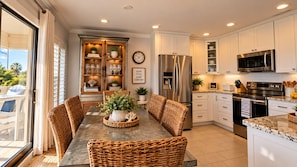 Una cocina moderna con muebles blancos, electrodomésticos de acero inoxidable y un comedor con sillas de mimbre y una mesa central.