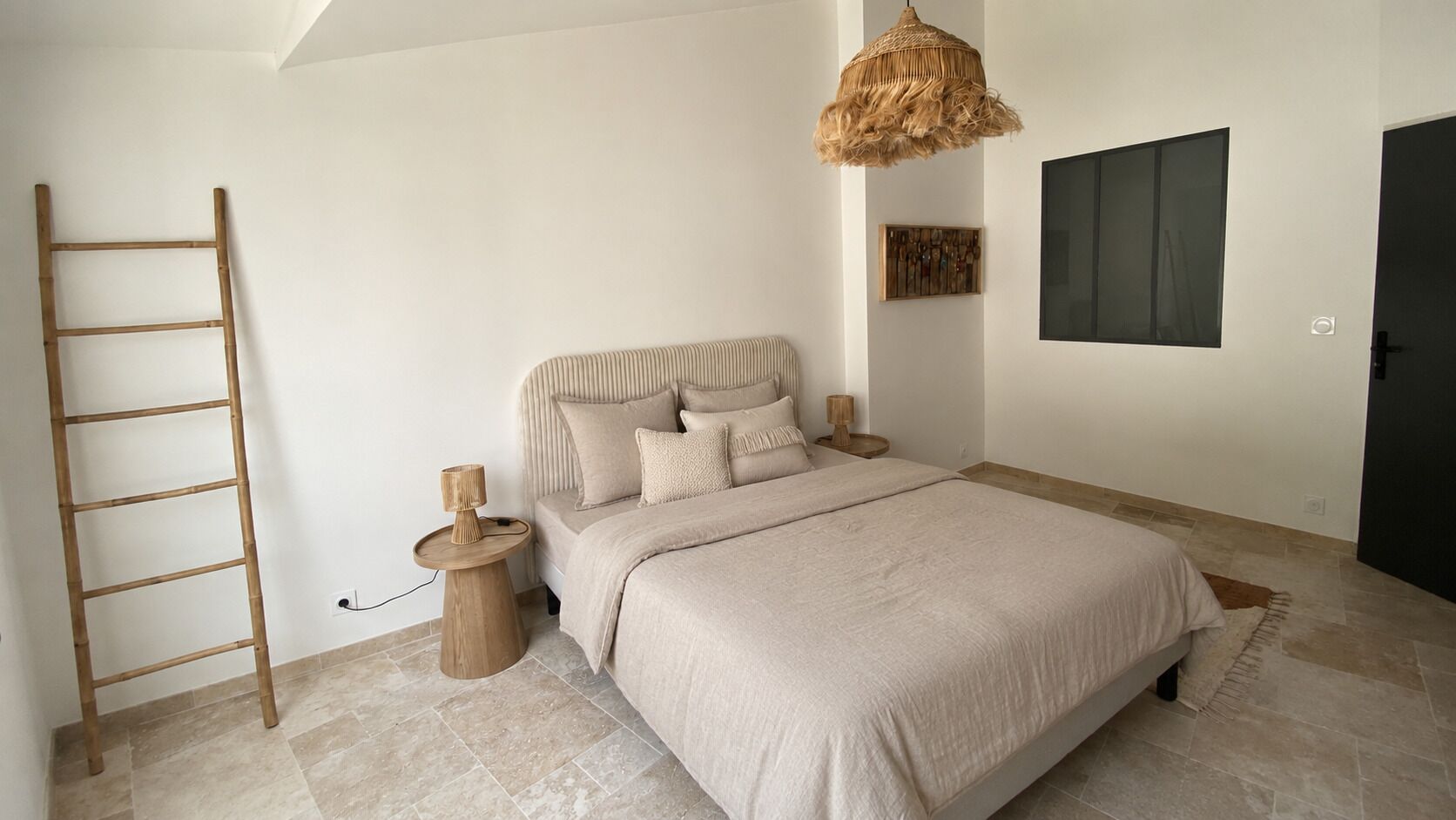 A bedroom with a bed, a ladder, a round wooden table with a lamp, a woven light fixture, and a framed artwork on the wall.