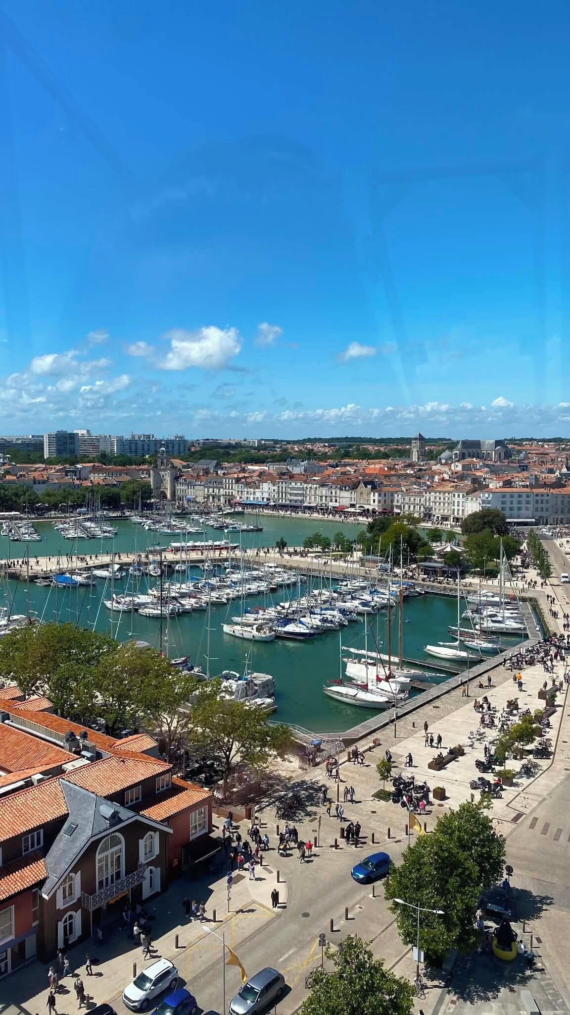 Een jachthaven met vele boten in de haven, een promenade langs het water en gebouwen met rode pannendaken.