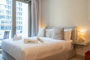 A hotel room with a bed, pillows, a bedside table with a lamp, and a window with curtains. - Marina Escape Steps from JBR Beach (Dubai)