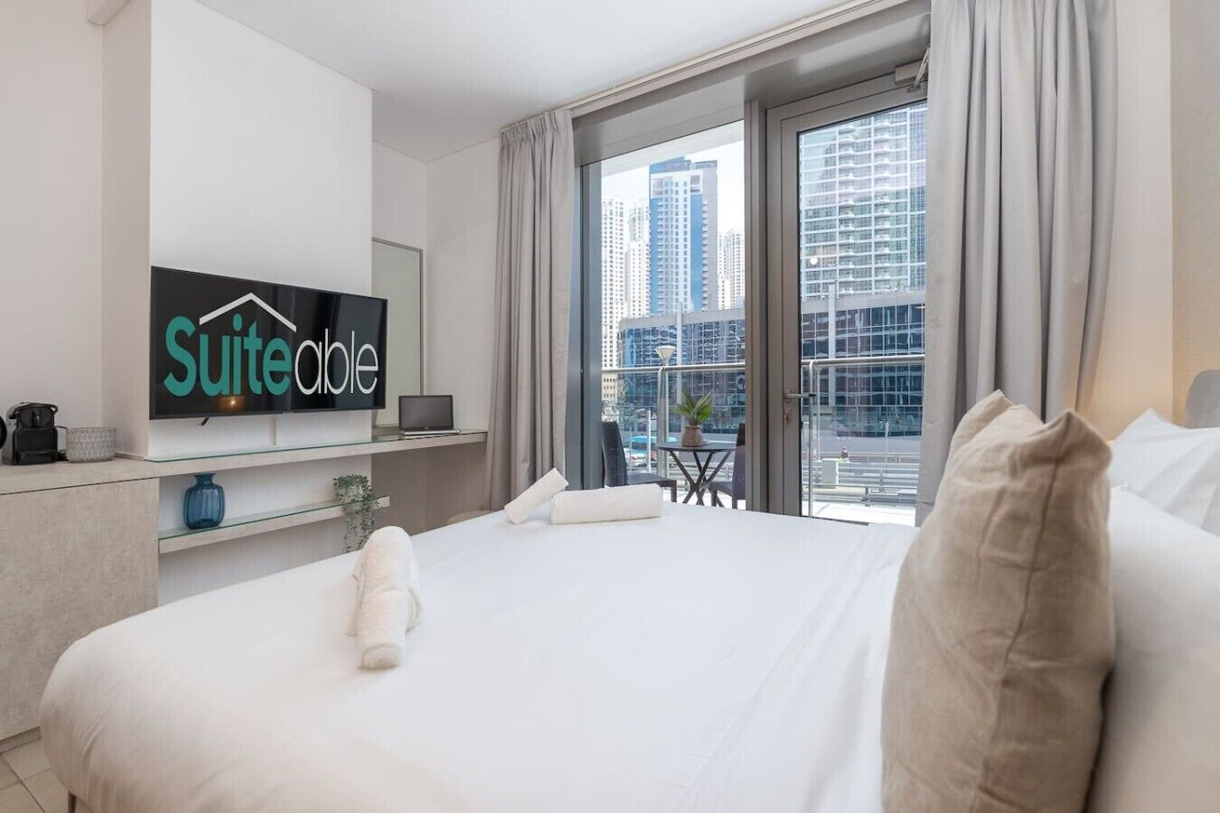 Ein Hotelzimmer mit Bett, einem Fernseher, der 'Suiteable' zeigt, einem Balkon mit Blick auf die Stadt und einer durch das Fenster sichtbaren Stadtansicht.