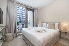 A hotel room with a bed, a desk, a chair, a small table, and a view of a building through the window. - Marina Escape Steps from JBR Beach (Dubai)