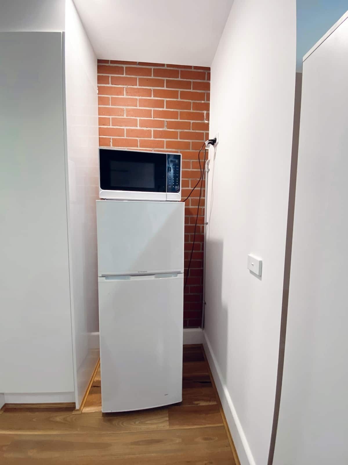 Een kleine keuken met een witte koelkast, een magnetron erboven en een bakstenen muur.