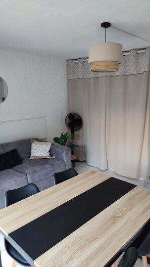 A modern living room with a grey sofa, a wooden dining table, and a ceiling fan. - One-bedroom apartment, sleeps 4, in Cap d'Agde, ground floor with garden, pets allowed (Agde)
