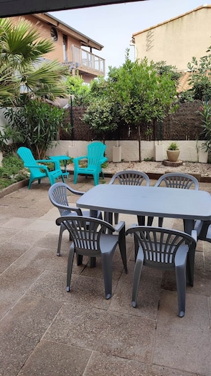 Een terras buiten met een tafel en stoelen, omringd door groen en een gebouw op de achtergrond.