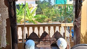 En balkon med gyngestol, et mønstret tæppe og udsigt til grønne områder og bygninger.