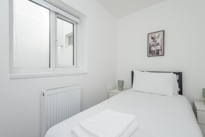A bedroom with a bed, a window, a radiator, a nightstand, a lamp, and a picture on the wall. - Hackney Central | London Fields & Broadway Market (London)