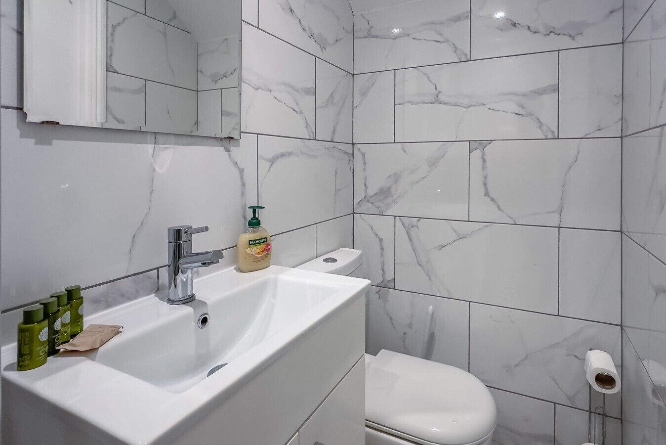 A bathroom with a white sink, mirror, and toilet, marble tiles, and a bottle of lotion.