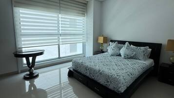 Un dormitorio con cama, mesitas de noche, una ventana con persianas y una pequeña mesa redonda.