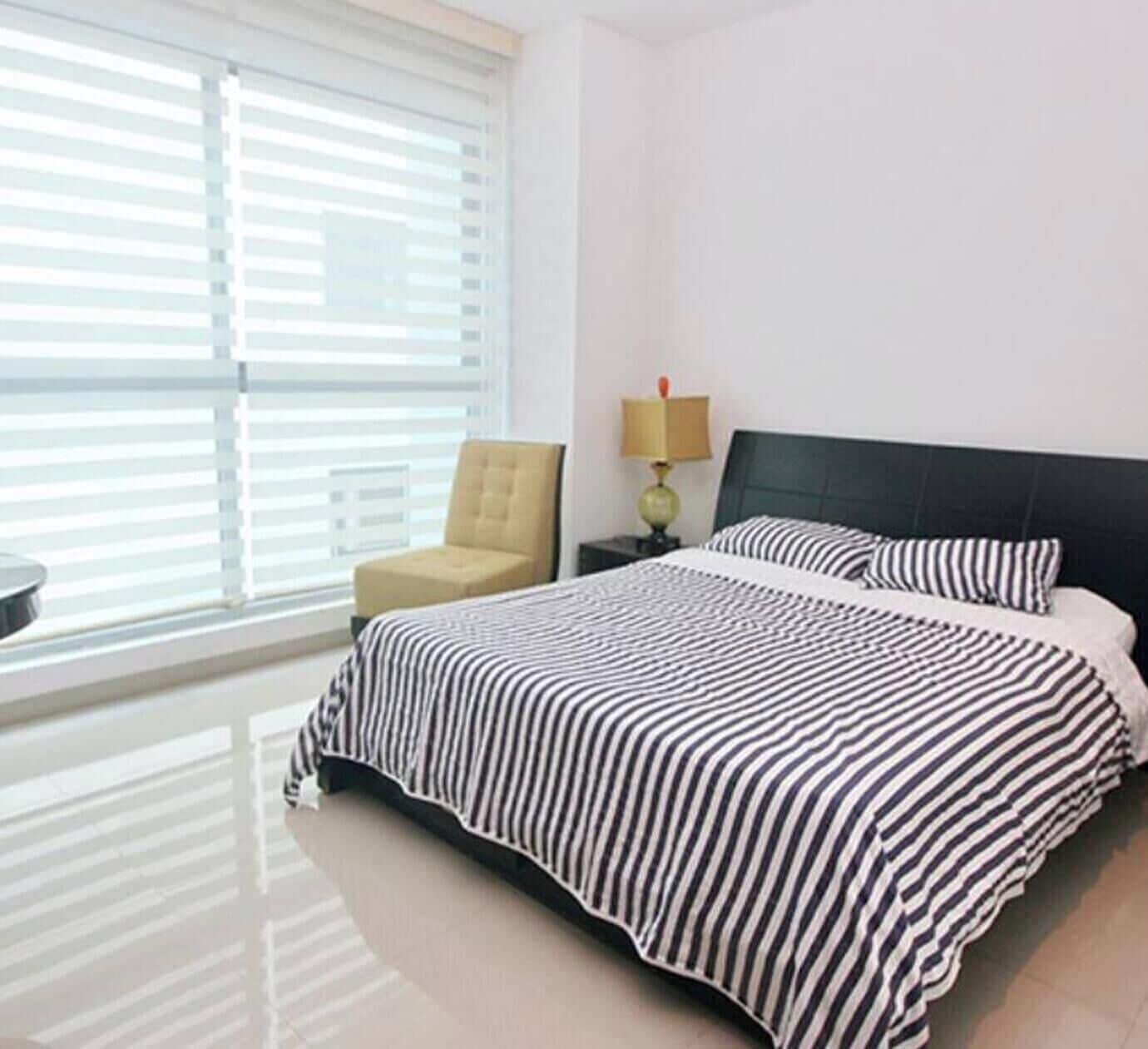 Una habitación de hotel con cama, una silla, una mesita de noche con lámpara y un ventanal con persianas.