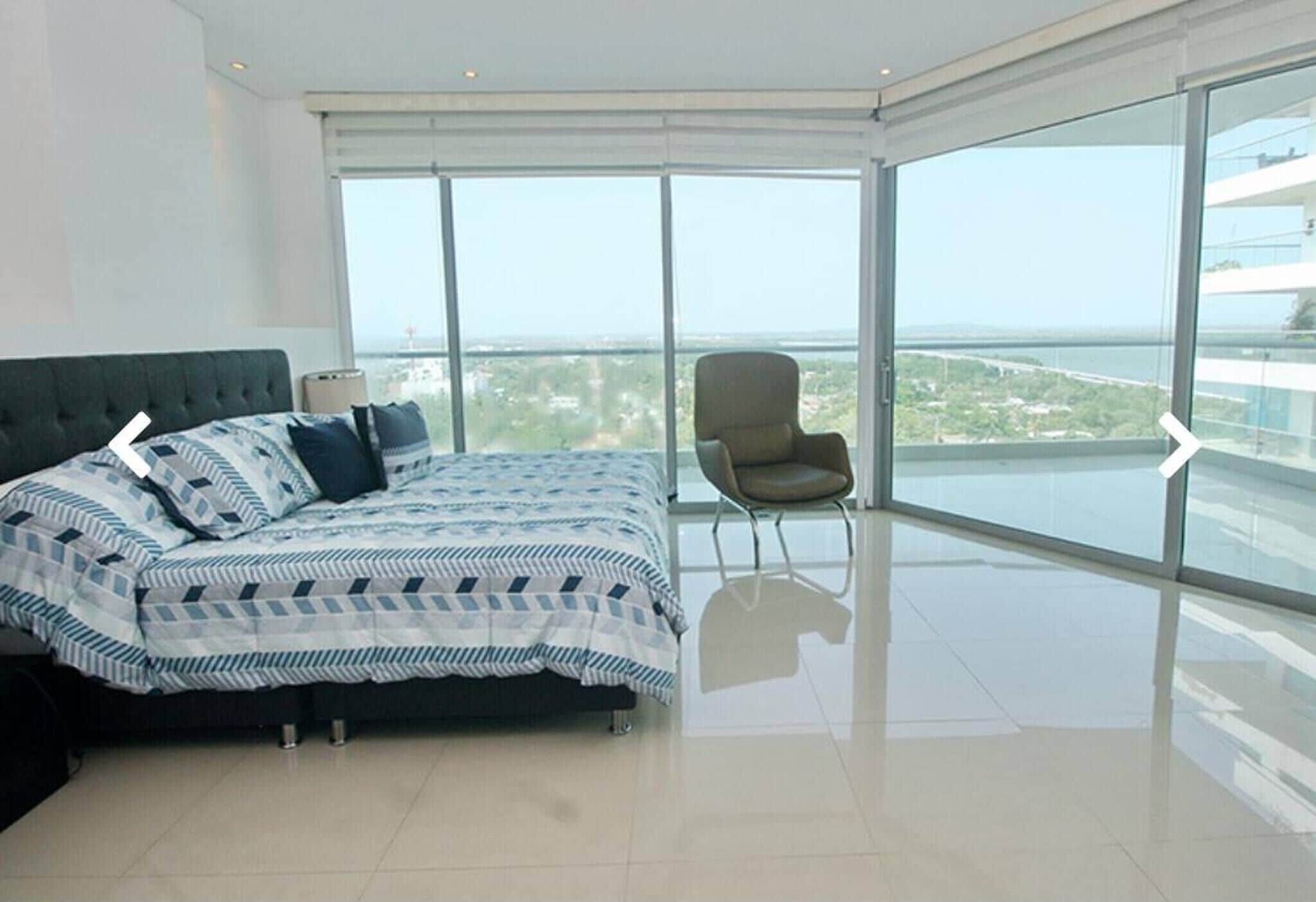 A modern bedroom with a large bed, a chair, and a balcony with a city view.