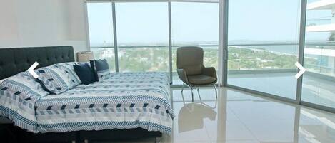 A modern bedroom with a large bed, a chair, and a balcony with a city view.