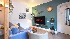A modern living room with a blue sofa, a white armchair, a coffee table, a flat-screen TV, and a wall-mounted shelf with a plant. - At the Bretzel Doré - Historic District (Colmar)