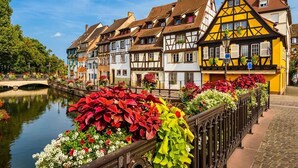 A row of traditional half-timbered houses along a canal with a bridge, surrounded by colorful flower beds. - At the Bretzel Doré - Historic District (Colmar)