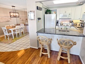 A kitchen with white cabinets, a refrigerator, a microwave, and a dining area with a table and chairs. - Oceanfront 2 Bed 2 Bath Condo with Pool! Braemar Towers 1804 (Ocean City)