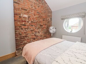 A bedroom with a brick wall, a round window, a bed with a white and grey patterned bedspread, and a white radiator. - Barn 1 (Sandbach)