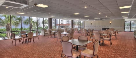 A large conference room with multiple tables and chairs, large windows with a view of palm trees, and a carpeted floor.