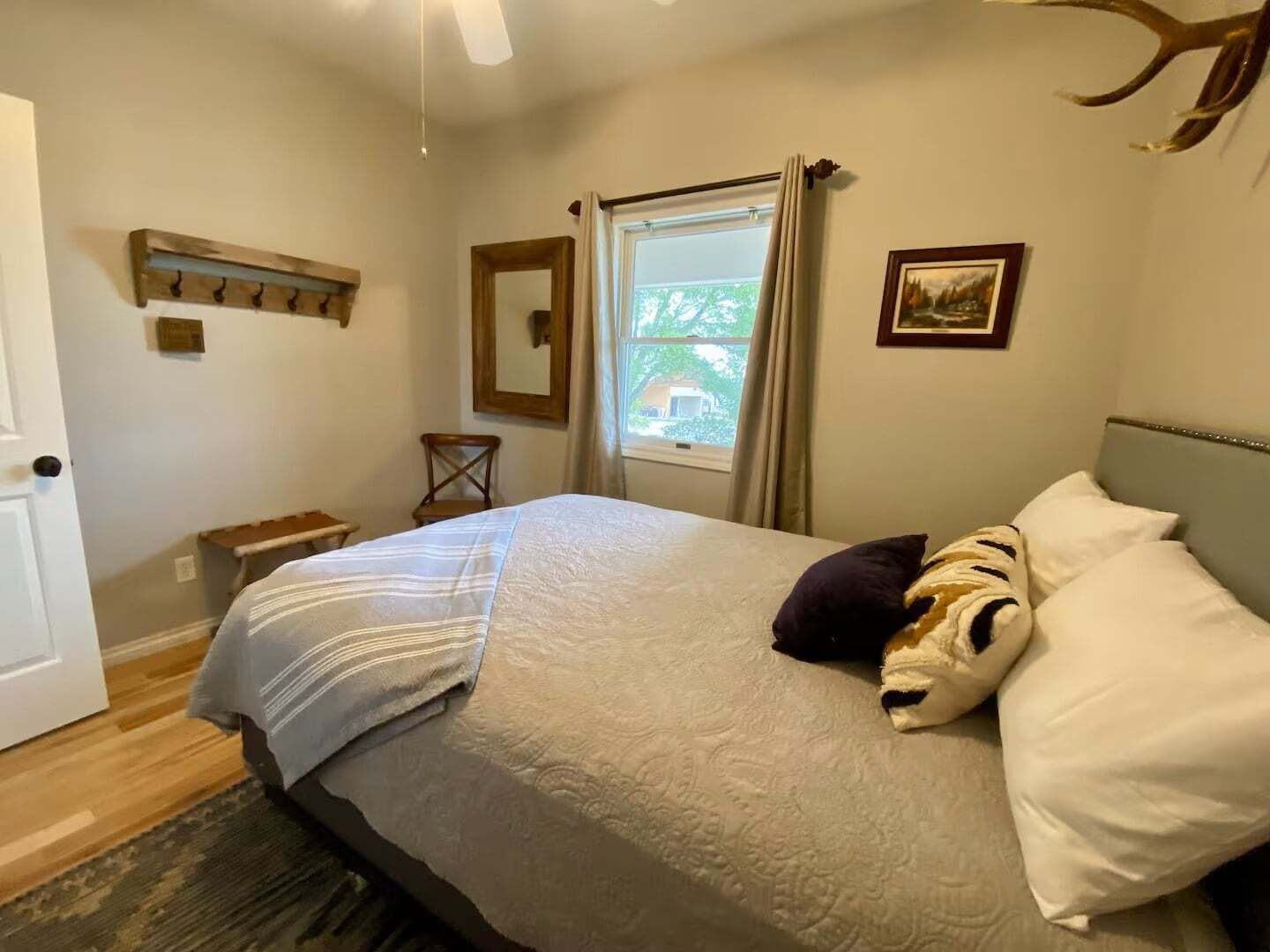 A bedroom with a bed, a window with curtains, a wooden headboard, a wall-mounted picture, and a ceiling fan.