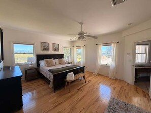 Un dormitorio con cama, mesitas de noche, ventilador de techo y ventanas con cortinas.
