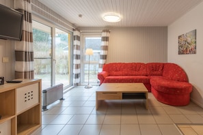 A living room with a red sofa, a wooden coffee table, a TV mounted on the wall, and a large window with curtains. - Skandinavischer Doppelbungalow mit Kaminofen im Ferienpark Mirow (Mirow)