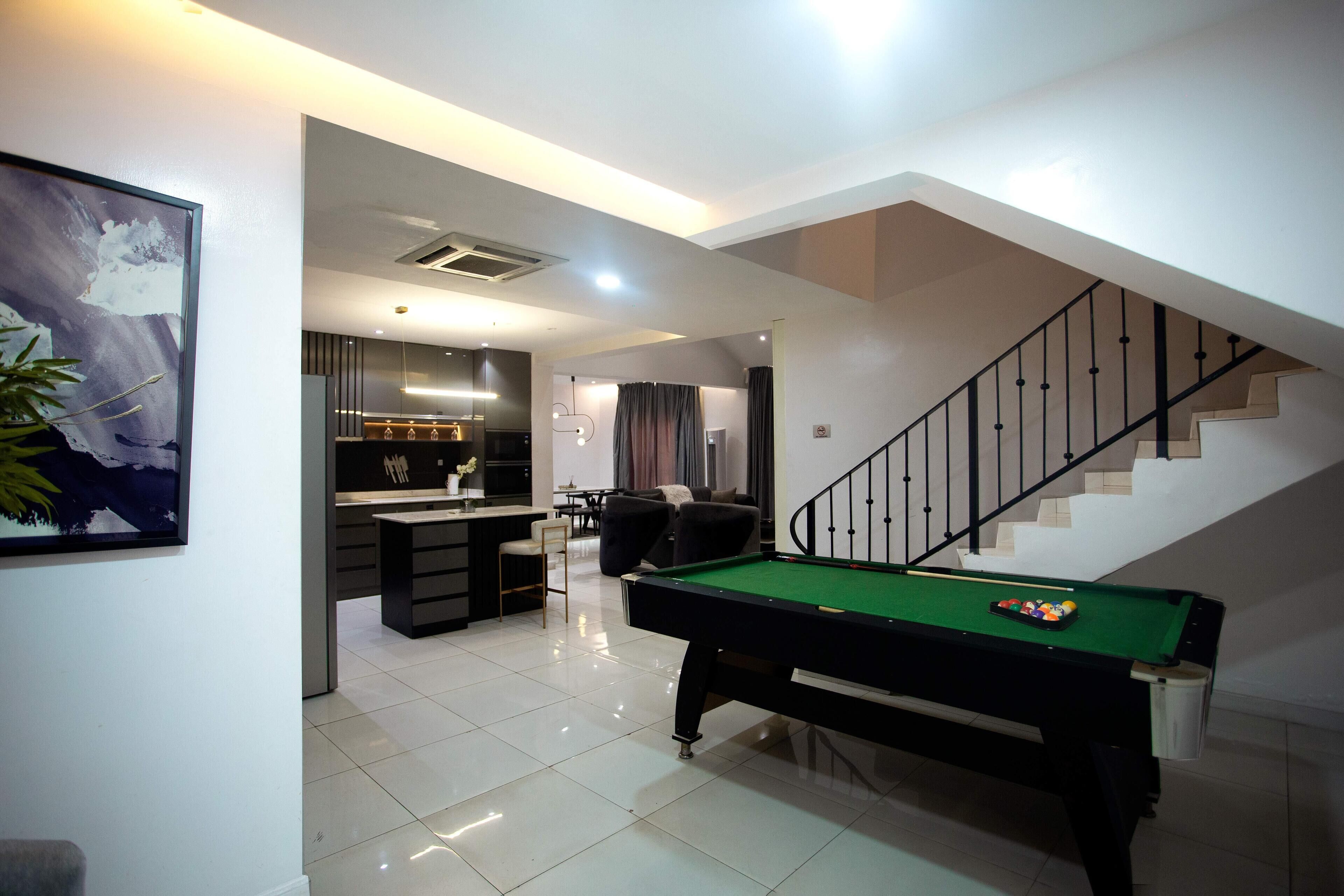 A modern living room with a pool table, a staircase, and a kitchen area.