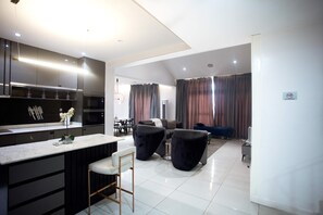 A modern kitchen with dark cabinetry and a central island, a dining area with a table and chairs, and a living area with a sofa and curtains. - DH Cadogan (Lekki)