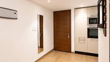 Um quarto com piso de madeira, porta, ar-condicionado fixado na parede e uma cozinheneta com micro-ondas e forno.